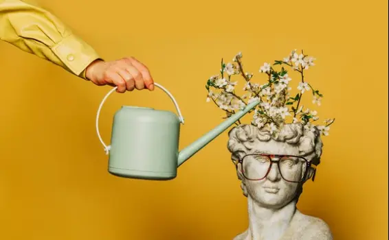 Mano regando un busto clásico con gafas del que brotan ramas en flor, metáfora del coaching transpersonal y el crecimiento interior.